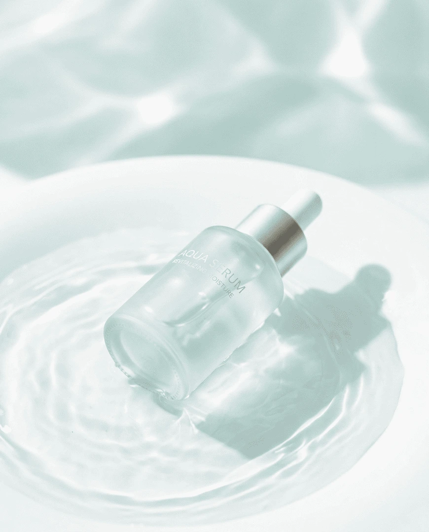 "A high-end commercial photography shot of a single, sleek frosted glass skincare serum bottle resting gently in a shallow pool of crystal-clear water"